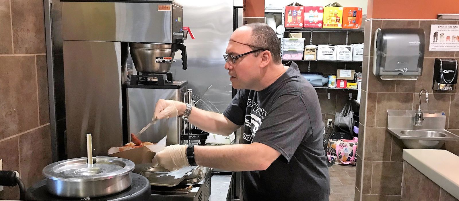LCS participant in a dark gray t-shirt is holding a ladle and scooping marinara sauce onto a food order for a customer